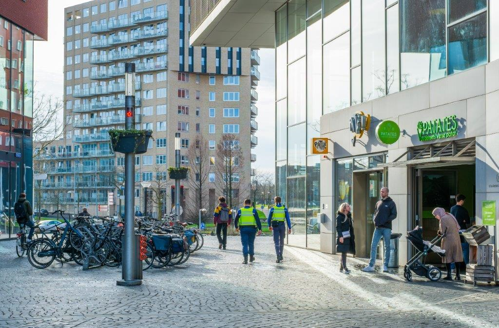  Een persoon met een blindengeleidestok en een persoon in een rolstoel, lopen door een winkelstraat. Op de voorgrond is een geparkeerde fiets zichtbaar. 